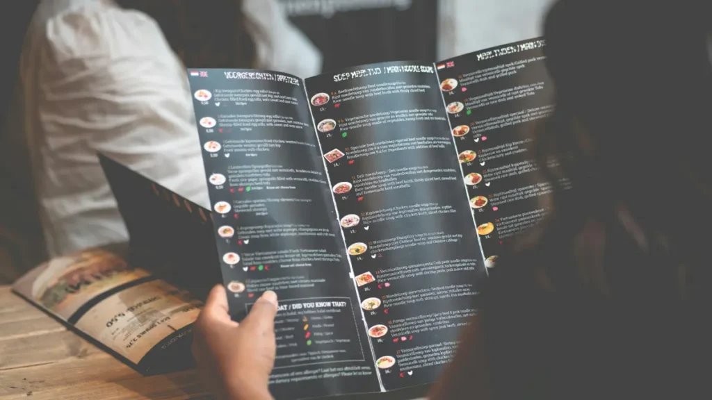 A person holding an open restaurant menu over a wooden table. The menu features images and descriptions of various dishes. Warm, inviting ambiance. Newks Menu Calories 